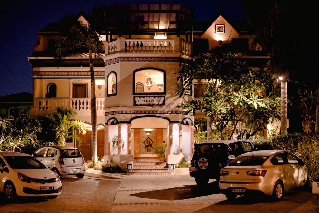 Modern automobiles parked on paved terrace with tall green plants near expensive residential house with glowing windows at night time